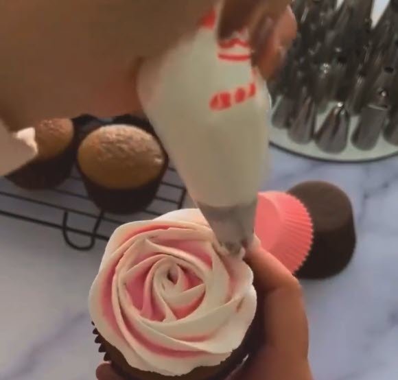 mão decorando cupcake de chocolate com bico de confeiteiro fazendo rosa degradê em tons de rosa e branco